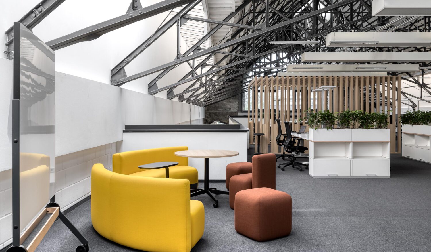 Office Köln: Moderne Büroumgebung mit gelben und braunen Sitzmöbeln, einem Tisch und einer offenen Raumstruktur mit sichtbaren Dachbalken.